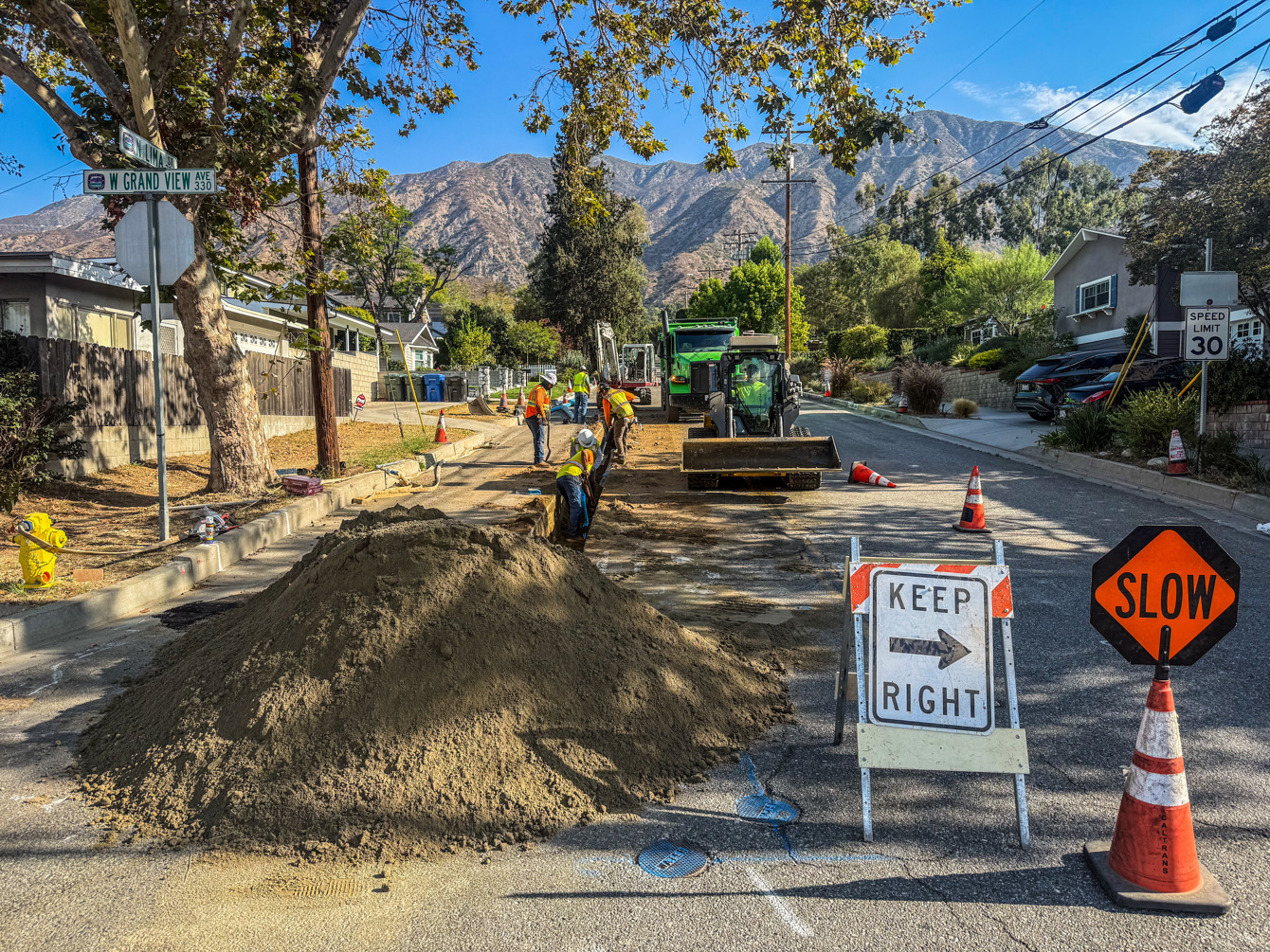 Crews trenching along North Lima Street behind Keep Right and Slow signs and traffic cones, with the San Gabriel Mountains in the background.